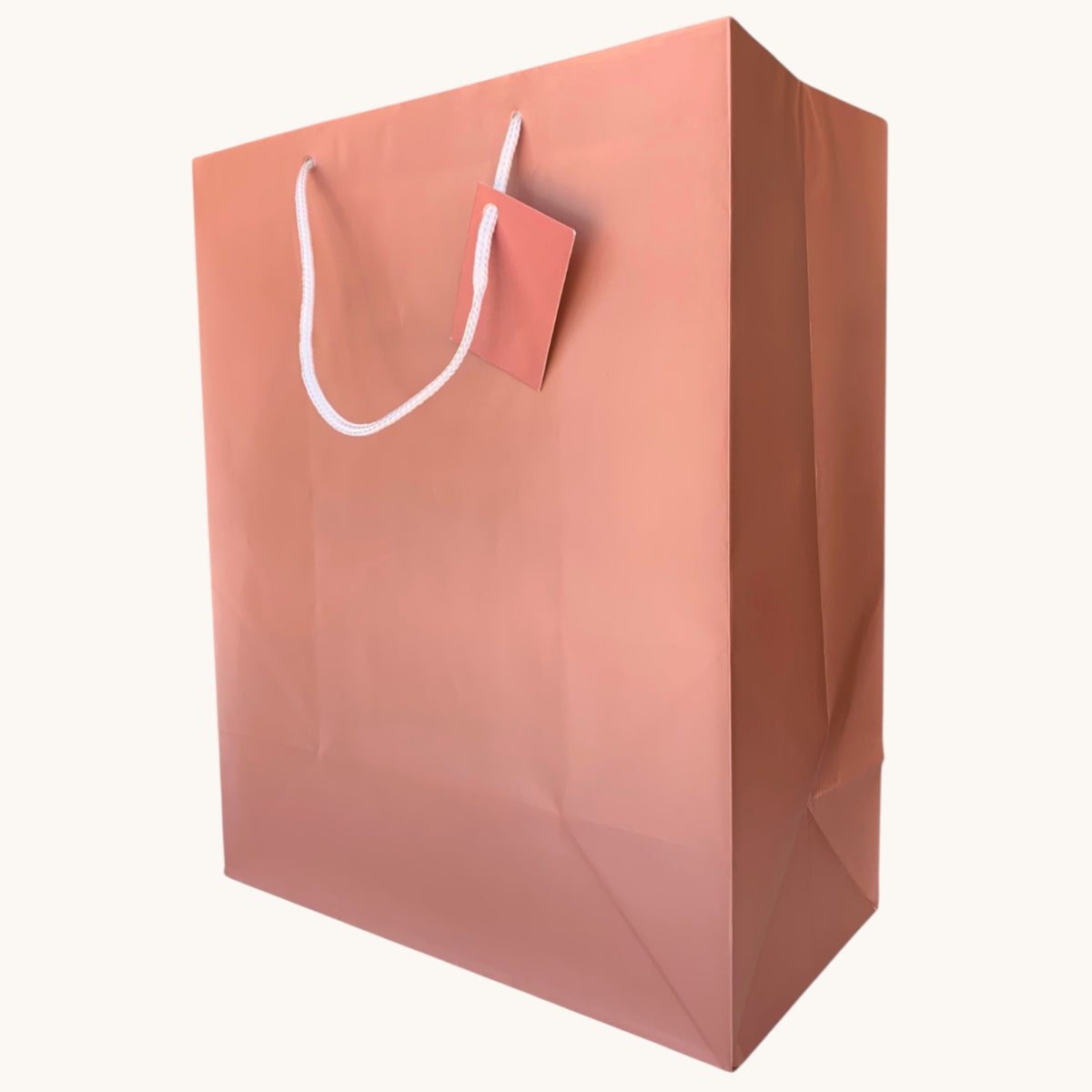 A large, empty rose gold paper gift bag in a dusty rose or muted pink colour, featuring white chord handles and a blank pink tag attached near the top, set against a plain off-white background.