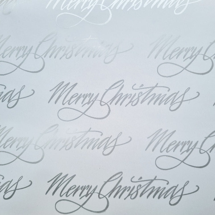 A close-up of white Christmas wrapping paper with metallic silver text that reads 'Merry Christmas' repeated across the surface.