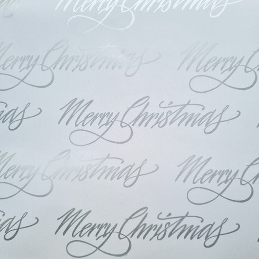 A close-up of white Christmas wrapping paper with metallic silver text that reads 'Merry Christmas' repeated across the surface.